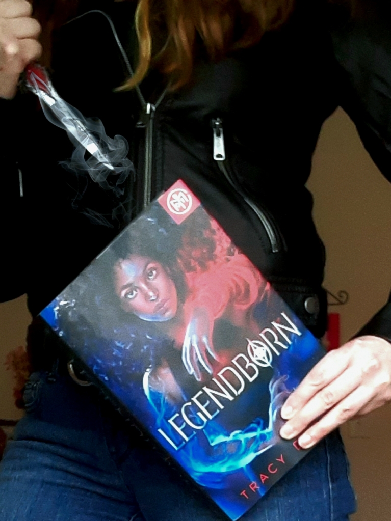 A white woman wearing a black leather jacket and blue jeans holds Legendborn by Tracy Deonn at her waist, and a prop dagger in her other hand above the book. The dagger is surrounded by silver smoke.